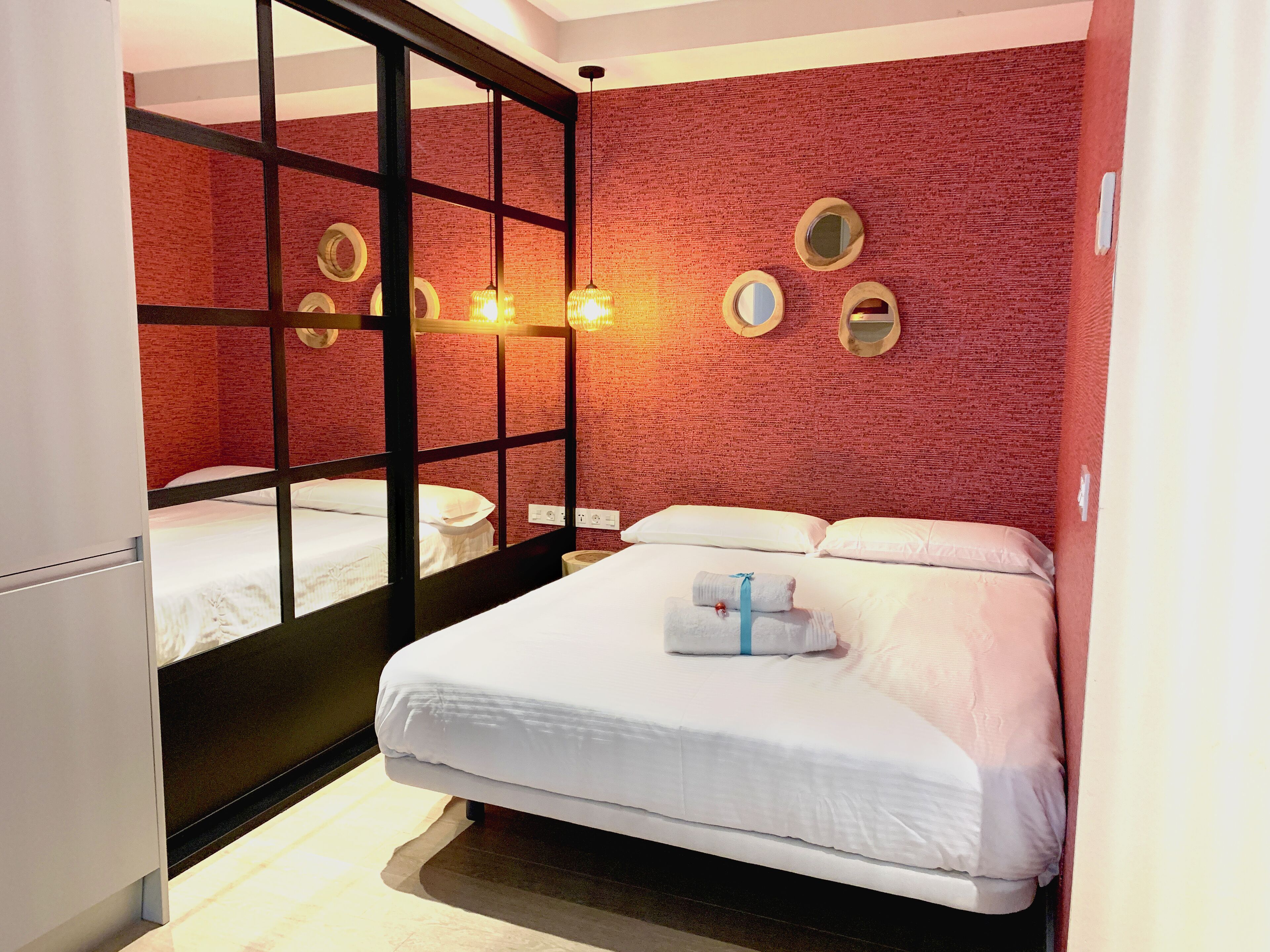 A hotel room with a mirrored wall, a bed with white linens, and a red textured wall.