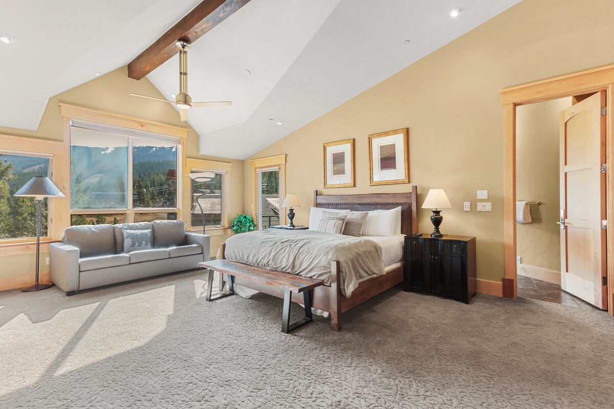 A spacious bedroom with a large bed, a sofa, and a bench. There are two windows with mountain views, a ceiling fan, and framed pictures on the wall.