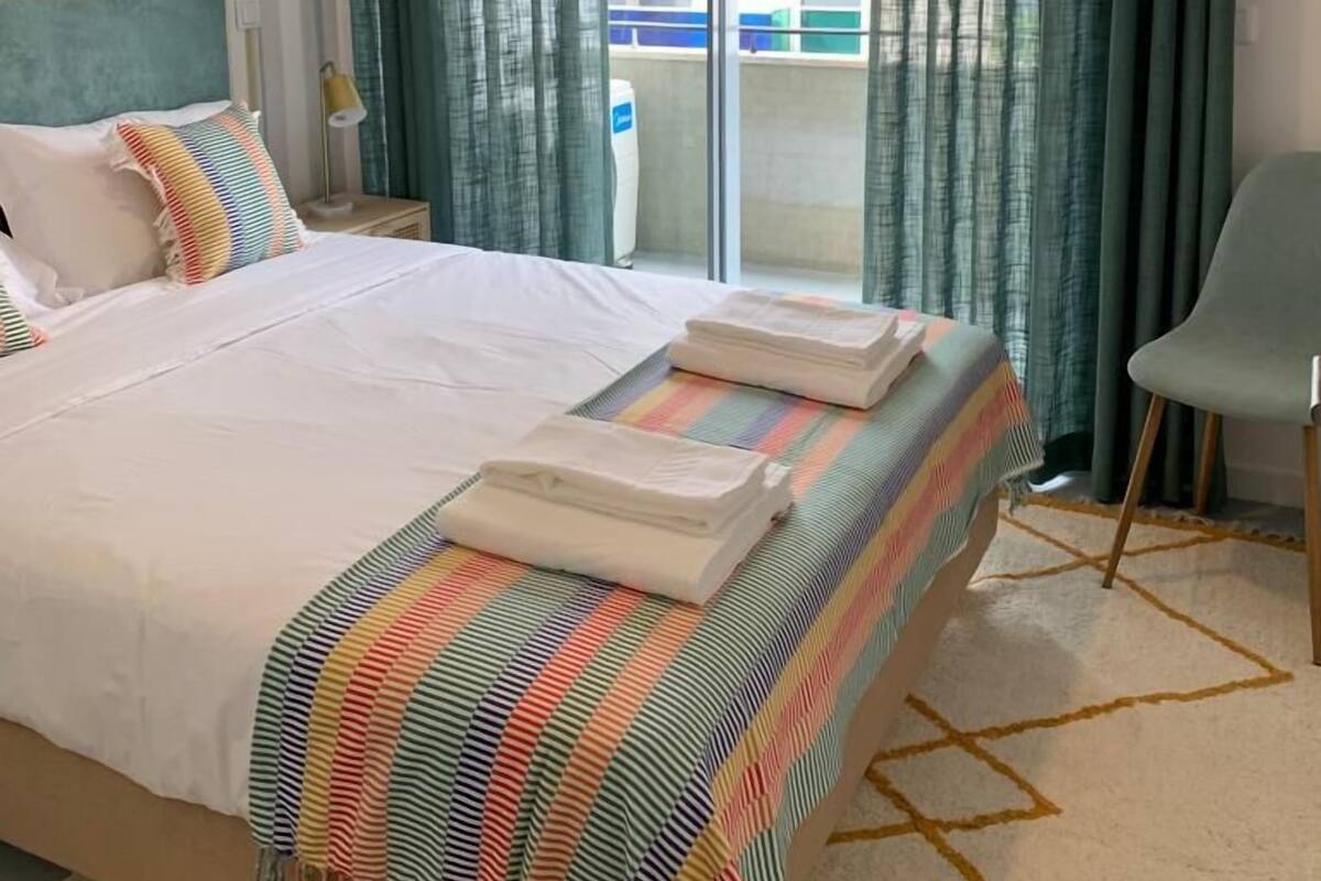 Une chambre d’hôtel avec un lit, une chaise, un balcon donnant sur des bâtiments aux couleurs vives et un appareil de climatisation.