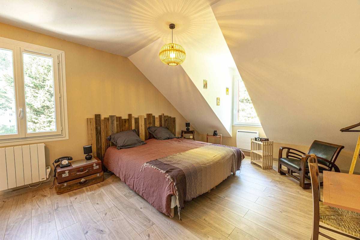 A bedroom with a sloped ceiling, a bed with a wooden headboard, a vintage suitcase, and a table with chairs.