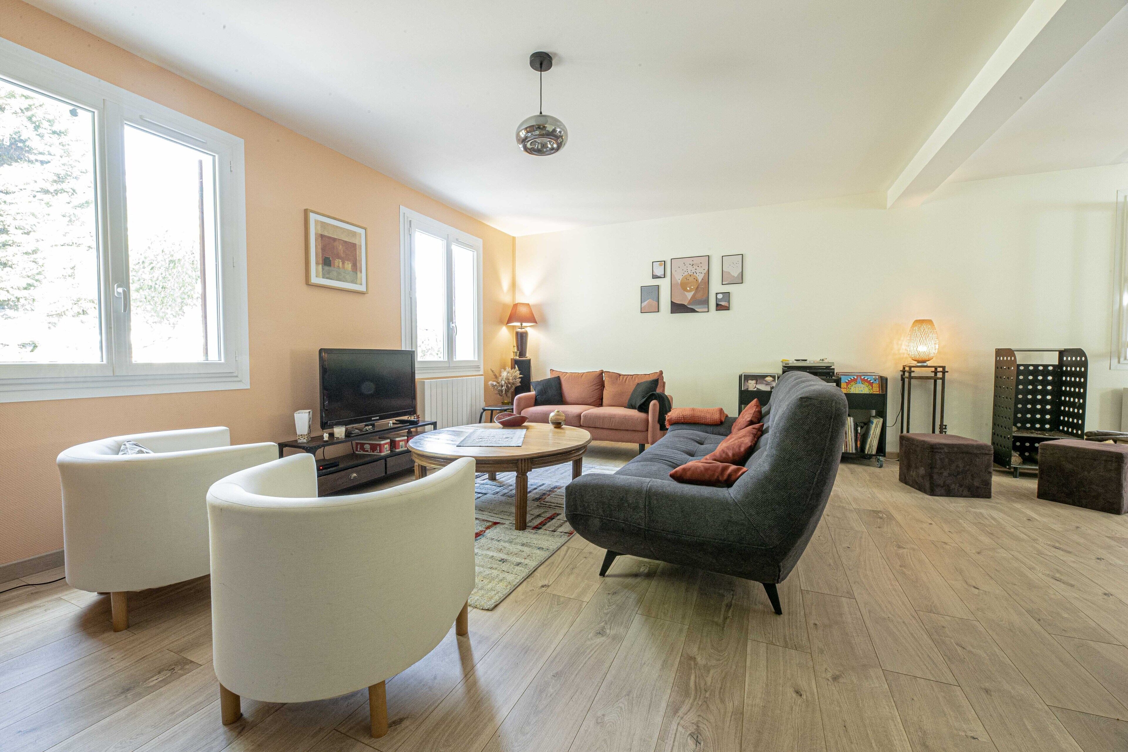 Un salon spacieux avec un canapé gris, des fauteuils blancs, une table basse et un téléviseur.