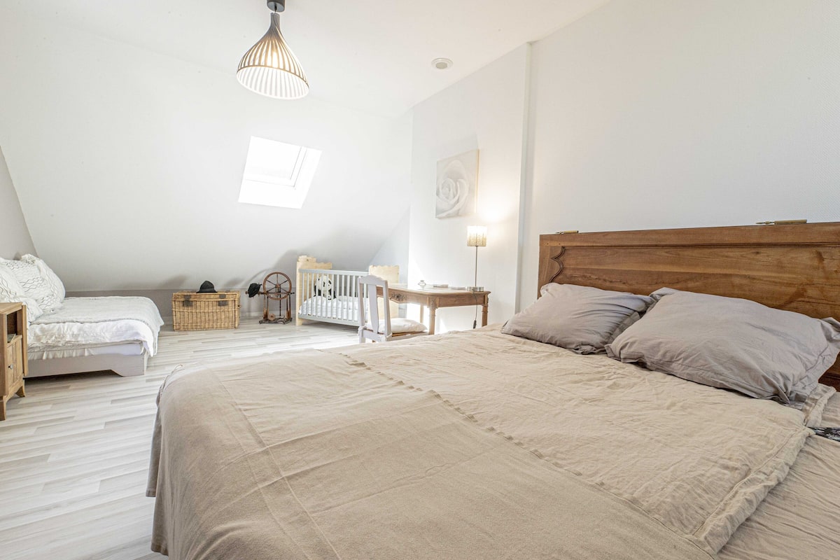A bedroom with a large bed, a crib, a wooden nightstand, a wicker basket, and a wall-mounted lamp.
