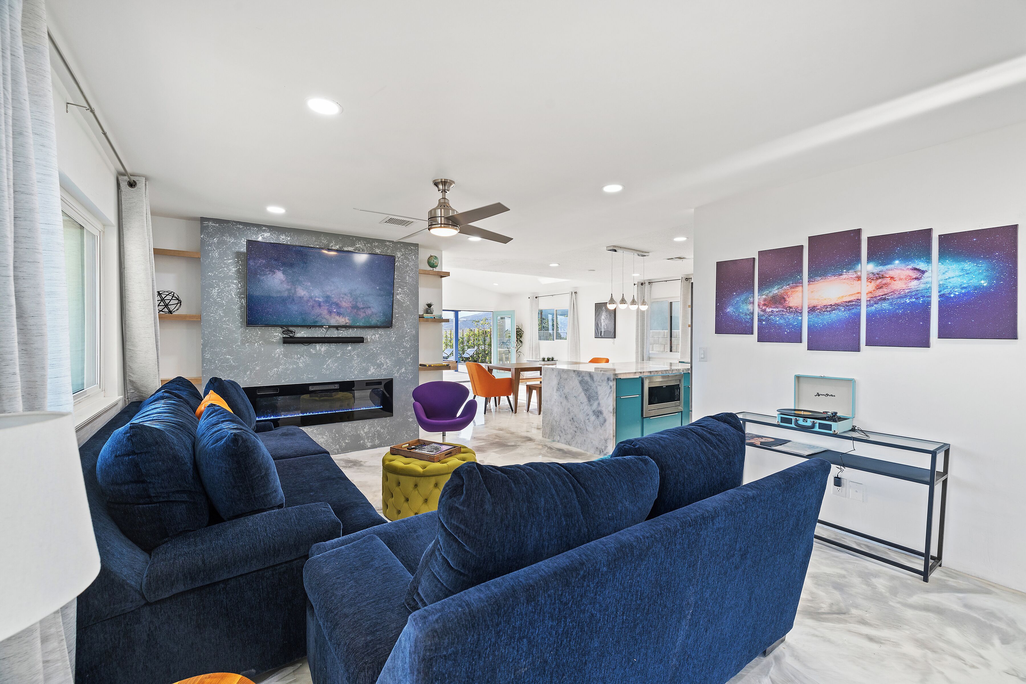 Una sala de estar moderna con un televisor de pantalla plana grande, sofás azules, un otoman amarillo y una cocina al fondo.