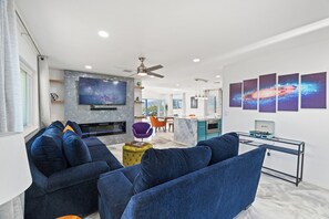 Una sala de estar moderna con un televisor de pantalla plana grande, sofás azules, un otoman amarillo y una cocina al fondo.