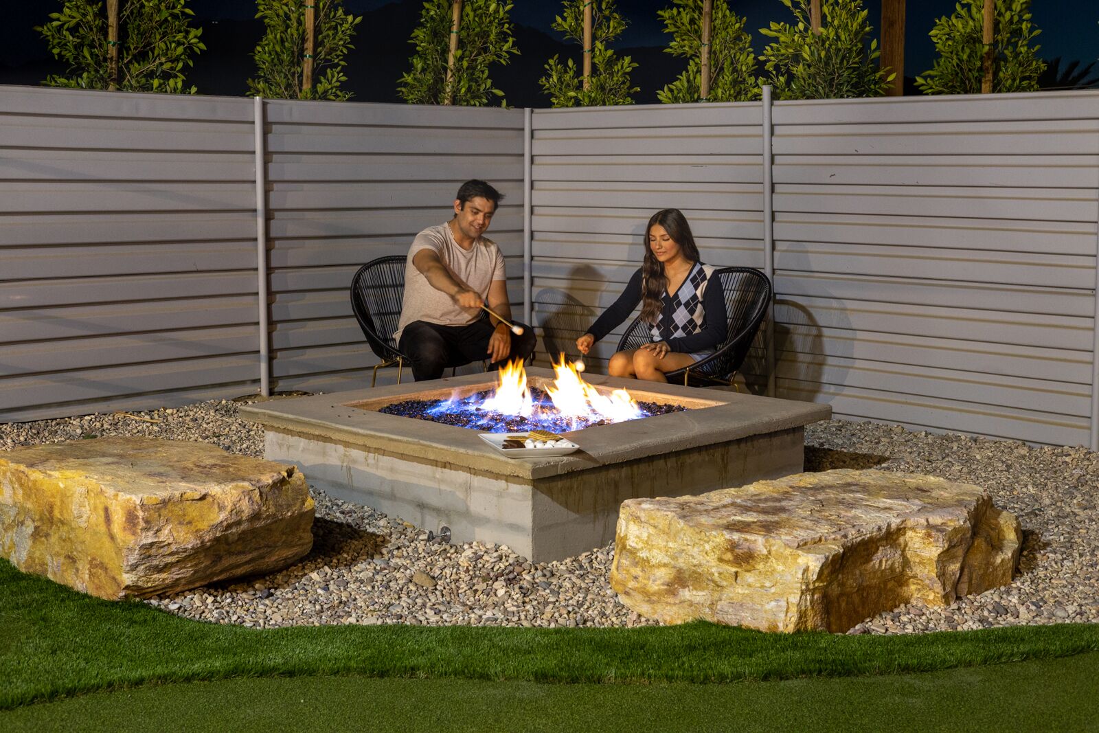 A fire pit with two people sitting on chairs, surrounded by rocks and a grassy area.