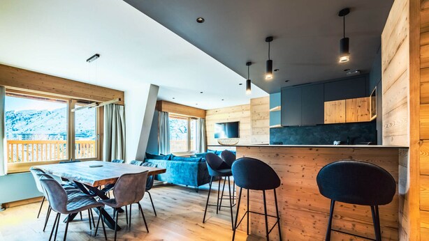 Een moderne keuken met houten details, een eethoek met een grote tafel en stoelen, en uitzicht op een besneeerd landschap door grote ramen.