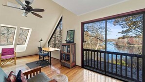 A spacious room with a large window overlooking a body of water, a ceiling fan, a wooden desk, a bookshelf, and a chair.