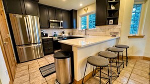 A modern kitchen with dark cabinets, stainless steel appliances, a large island with bar stools, and a window with a view of trees.