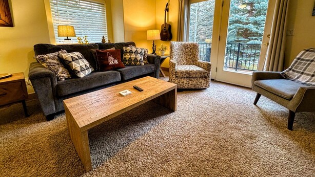 A cozy living room with a sofa, armchair, and coffee table, a guitar, and a view of trees through large windows.