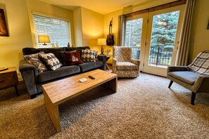 Una sala de estar acogedora con sofá, sillón y mesa de centro, una guitarra y vistas a árboles a través de amplios ventanales.