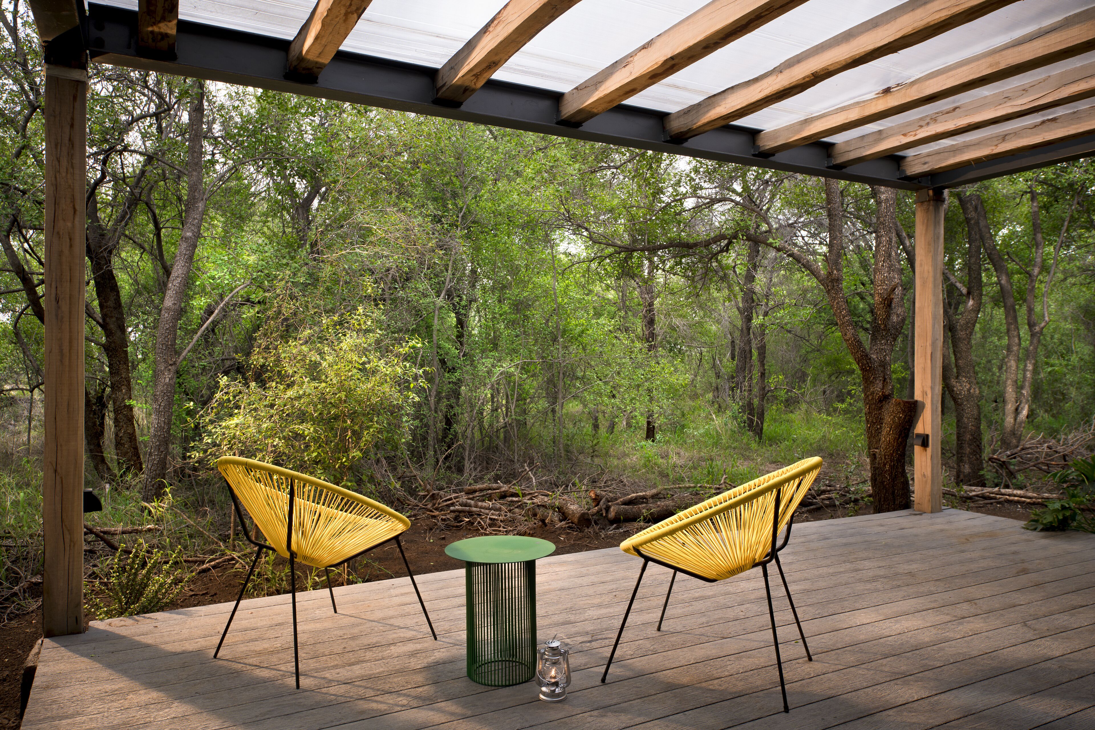 An outdoor wooden deck with two yellow chairs and a green table, surrounded by trees.