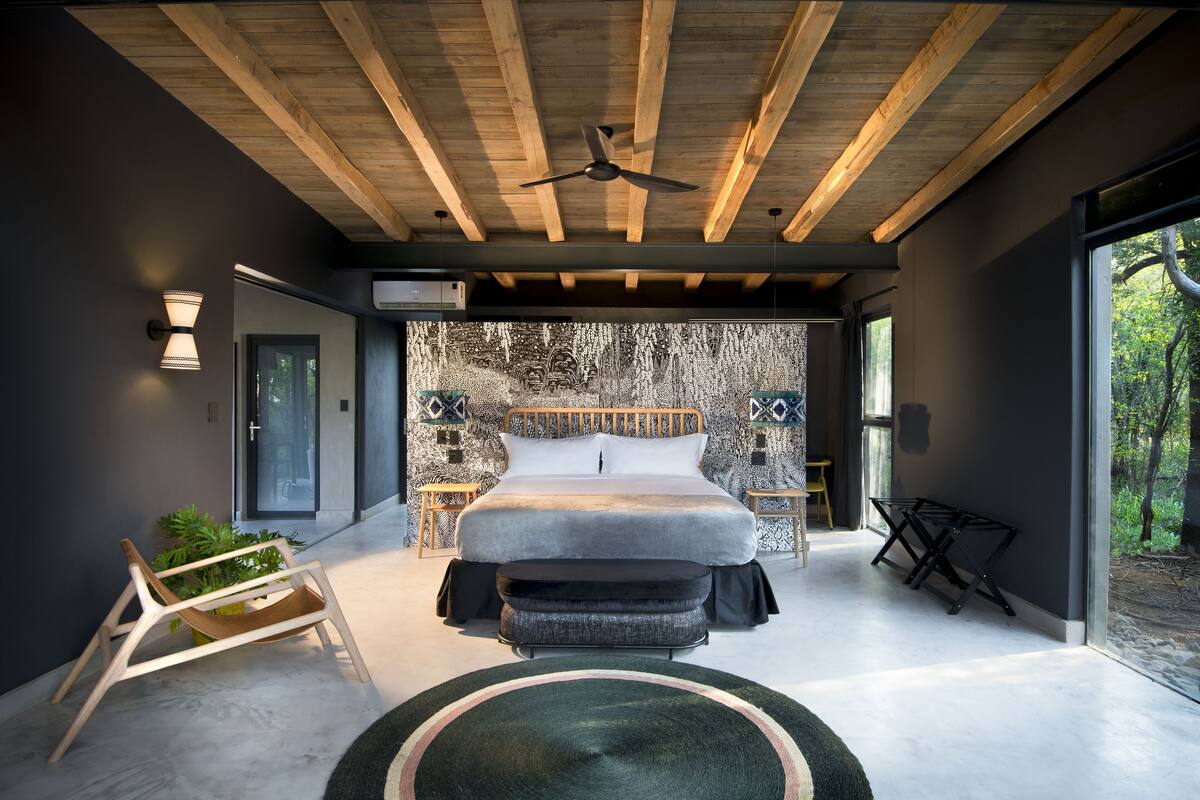 Une chambre moderne avec un grand lit, des poutres en bois au plafond, un ventilateur de plafond et une vue sur l’extérieur grâce à une porte vitrée.