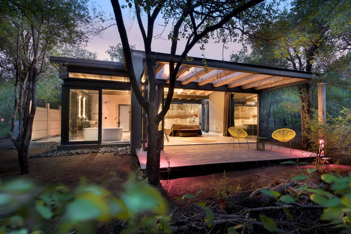 Une maison moderne avec une façade en verre, une terrasse en bois et un espace détente avec des chaises jaunes.
