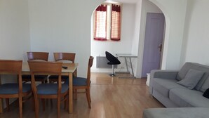 A living room with a dining area, a grey sofa, wooden chairs, a black chair, and a window with curtains. - 2 Pièces 50m² Ensoleillés + 2 Terrasses BBC (Menton)
