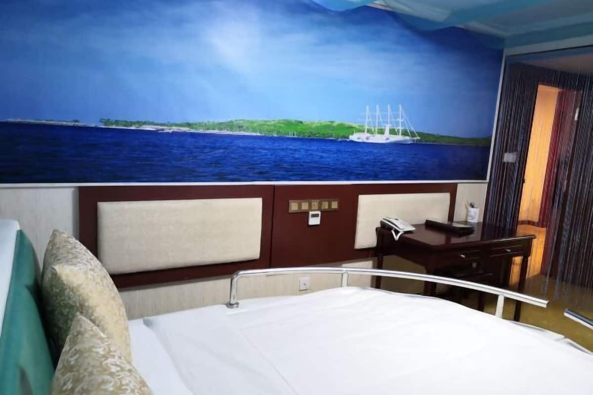 A hotel room with a bed, a desk, and a view of the ocean with a sailboat.