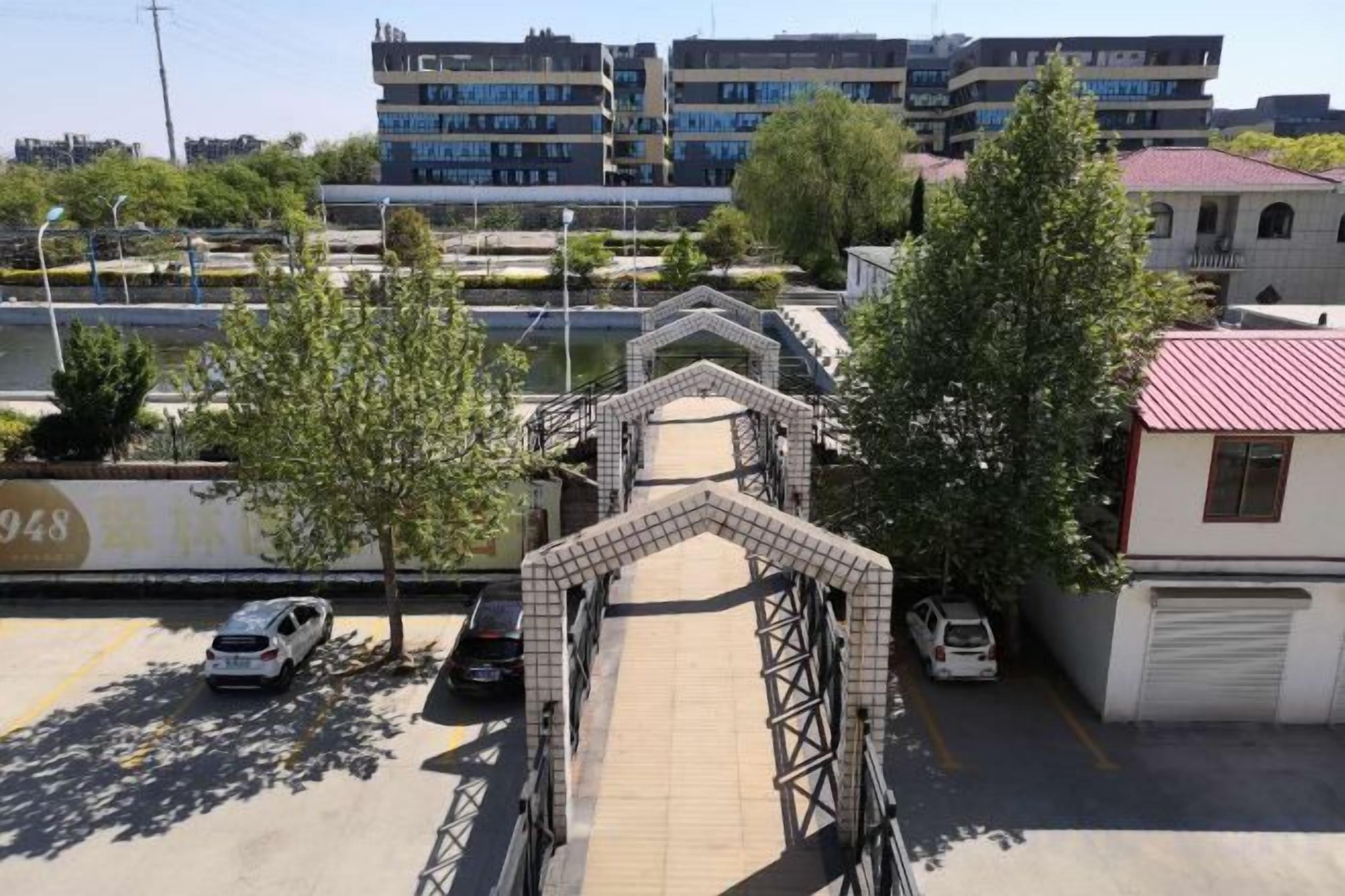 A pedestrian bridge with a metal framework, a parking area with cars, and buildings in the background.