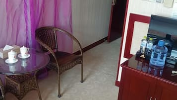 Una habitación pequeña con una mesa redonda de vidrio, sillas de mimbre, un armario rojo con botellas de agua y una cortina púrpura.