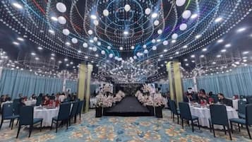 A banquet hall with tables arranged for dining, a stage at the front, and a ceiling adorned with hanging lights.