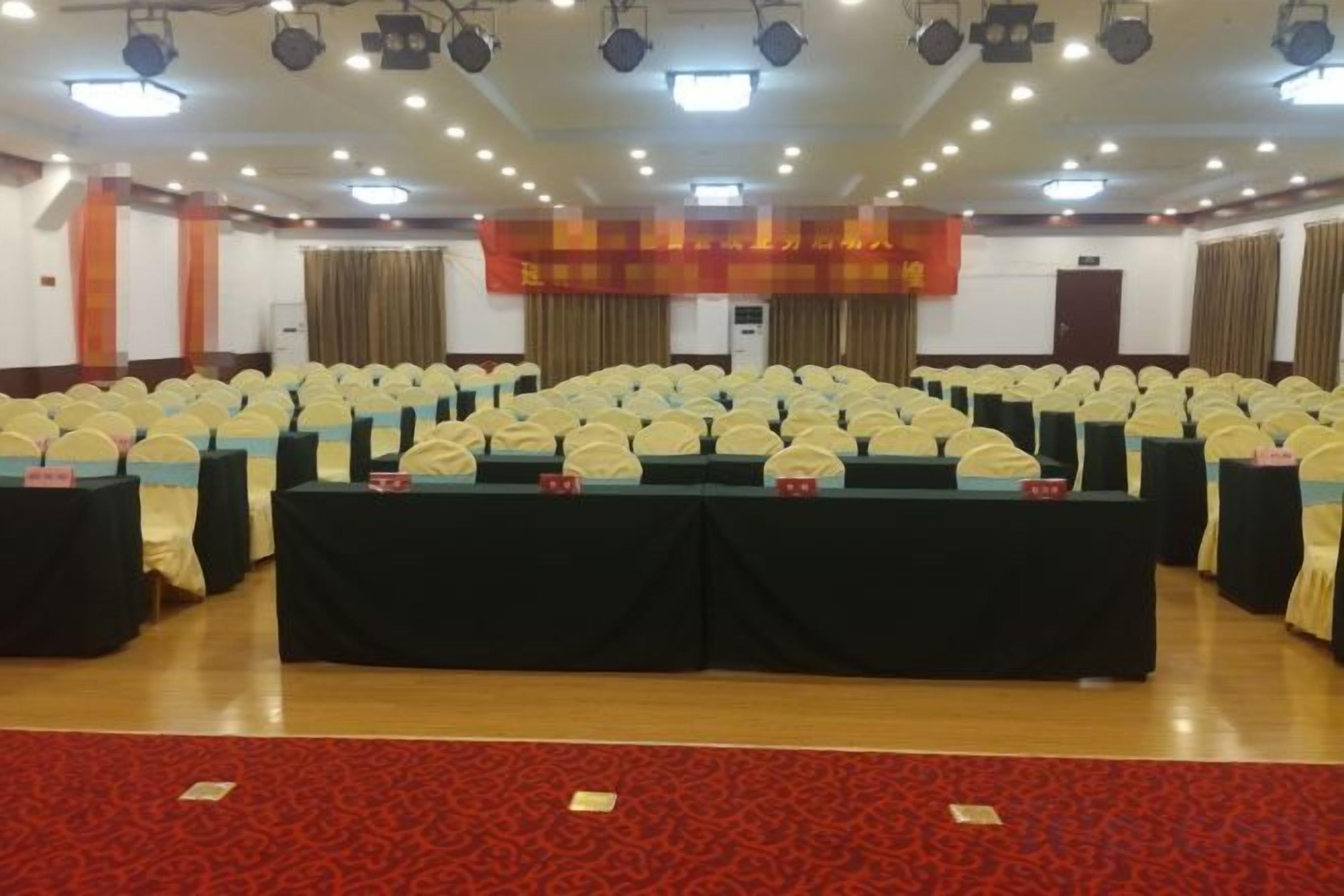 Un salón de conferencias con filas de sillas y mesas, una alfombra roja y un cartel en la pared.