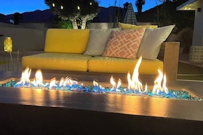 Ein Feuerstelle mit blauen Glassteinchen, ein gelbes Sofa mit gemusterten Kissen und ein dekorativer Laterne in einer Terrassensituation.