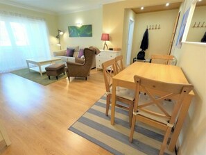 A living room with a sofa, armchair, coffee table, and a dining table with chairs. There is a lamp, a coat rack, and a painting on the wall.