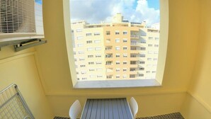 A balcony with a table and chairs, a window with a view of a building, and a metal railing. - Carnaxide, modern T1 apartment w/ balcony, Lisbon (Carnaxide)