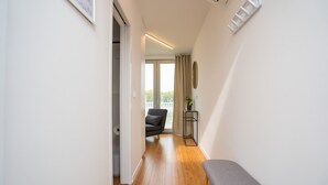 Un pasillo estrecho con suelo de madera, un banco gris, una silla negra y una pequeña mesa con una planta.