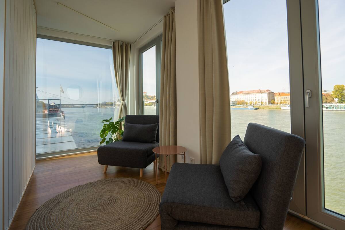 A modern room with a large window, two grey armchairs, a small round table, and a woven rug.
