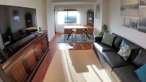 A living room with a TV, a dining area with a table and chairs, a sofa, and a large window. - Lisbon, T2 apartment in condominium, Linda-a-Velha (Linda a Velha)
