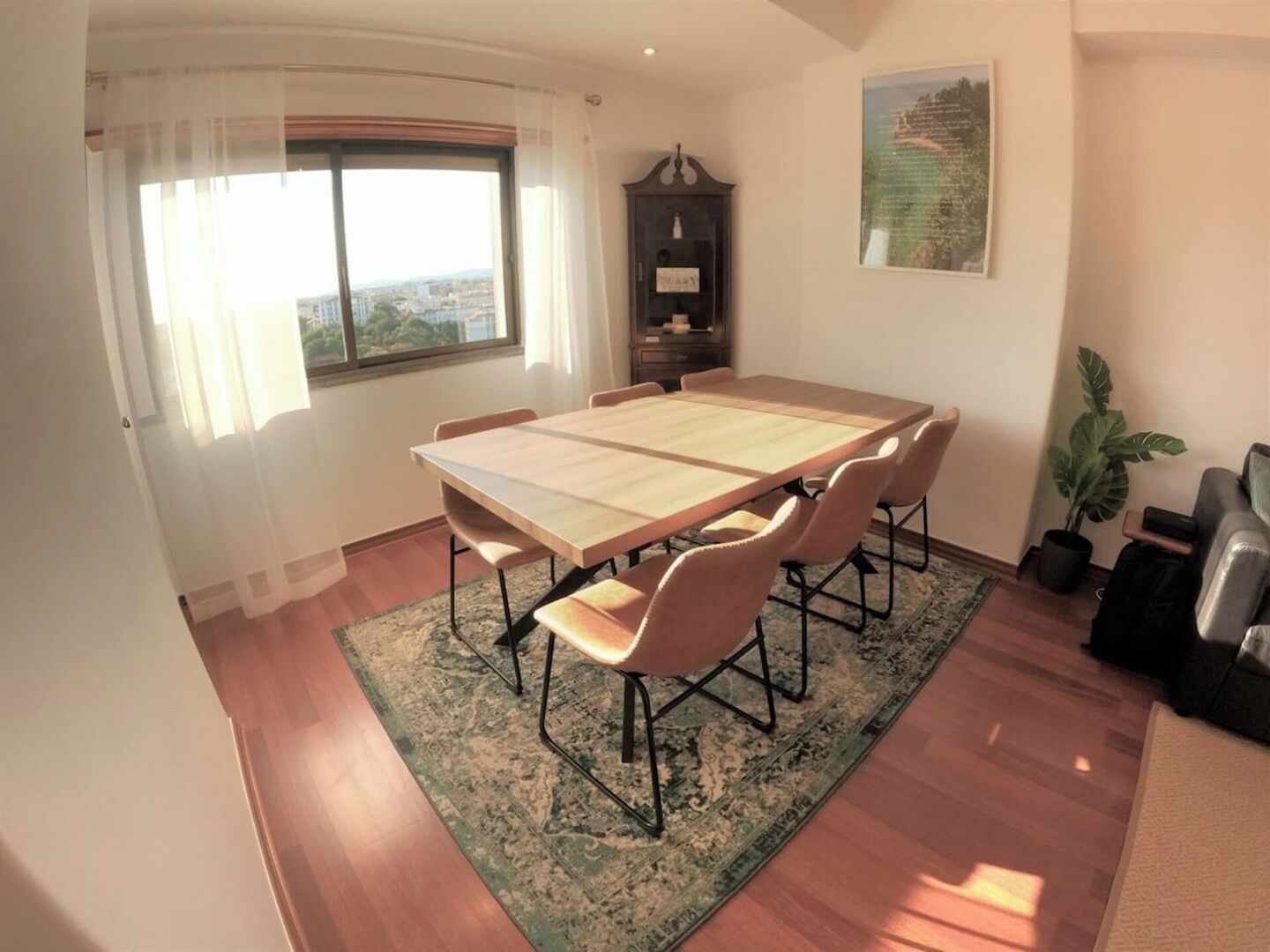 Ein Esszimmer mit großem Tisch, Stühlen, einem Schrank, einer Topfpflanze und Blick durch ein Fenster.
