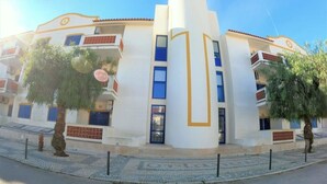 Un edificio moderno con una torre central, balcones y un área pavimentada con árboles.
