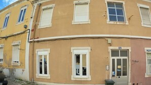 A two-story building with a yellow facade, white window frames, and a green trash bin in front. - Lisbon T2 apartment in typical villa, Sapadores (Lisboa ciudad)