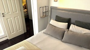 En pænt redt seng med puder, et sengebord med en lampe, og en dør der fører til et badeværelse.