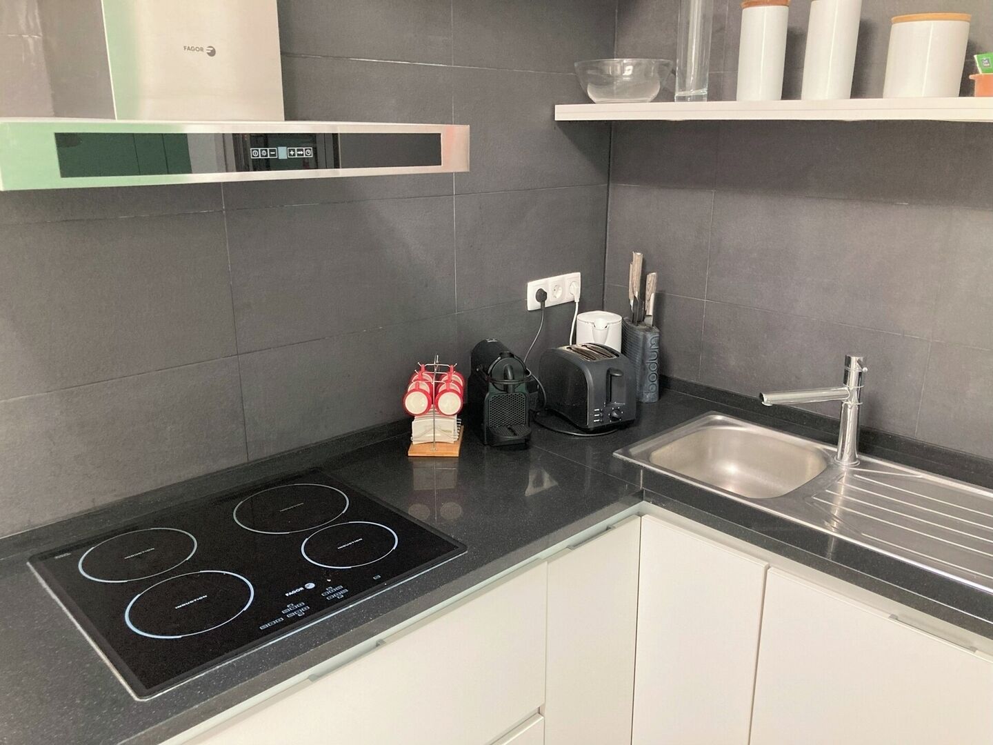 Una cocina moderna con una placa de inducción negra, horno empotrado, un lavamanos de acero inoxidable y un armario blanco.