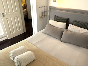 A neatly made bed with pillows and a rolled towel, a bedside table with a lamp, and a door leading to another room.