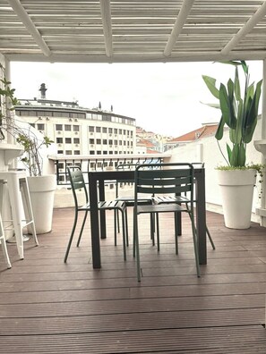 Un espace extérieur abrité, aménagé avec une table et des chaises, des plantes en pot et offrant une vue sur les bâtiments en arrière-plan.
