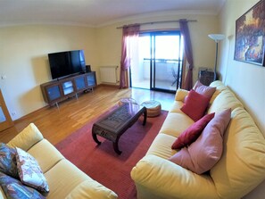 Un salon comprenant un canapĂ©, une table basse, un tĂ©lĂ©viseur posĂ© sur un meuble et une porte-fenĂȘtre coulissante donnant sur un balcon.