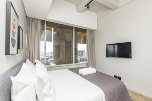A modern hotel room with a large bed, a flat-screen TV, and a city view through a large window.