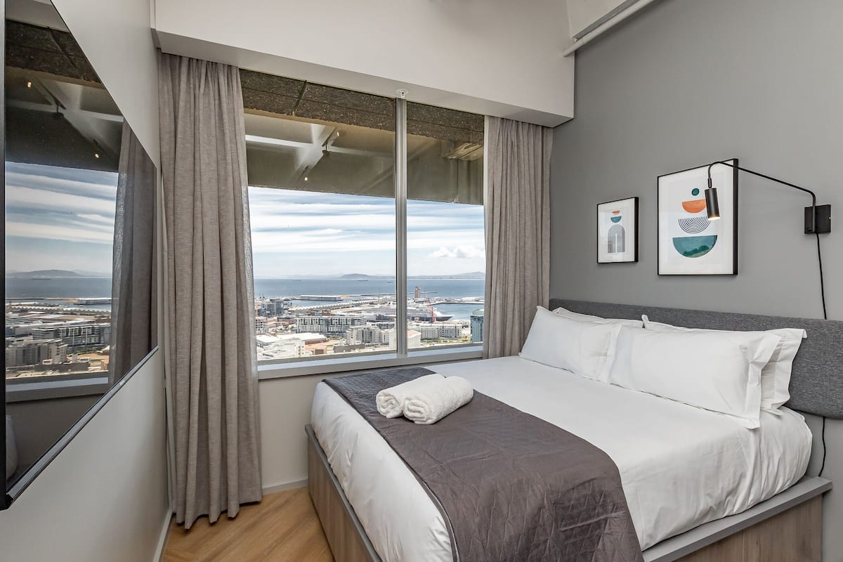 A modern hotel room with a large bed, a city view, and artwork on the wall.