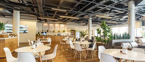 A modern office space with round tables and white chairs, a kitchen area, and large windows allowing natural light.