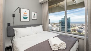 A modern hotel room with a large bed, a city view, and artwork on the wall.
