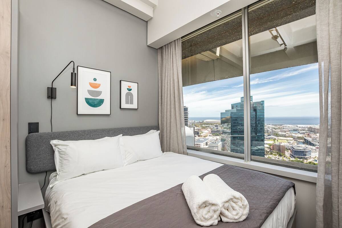 A modern hotel room with a large bed, a city view, and artwork on the wall.