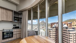 A modern kitchen with a round wooden table, large windows with a city view, and a view of a mountain.