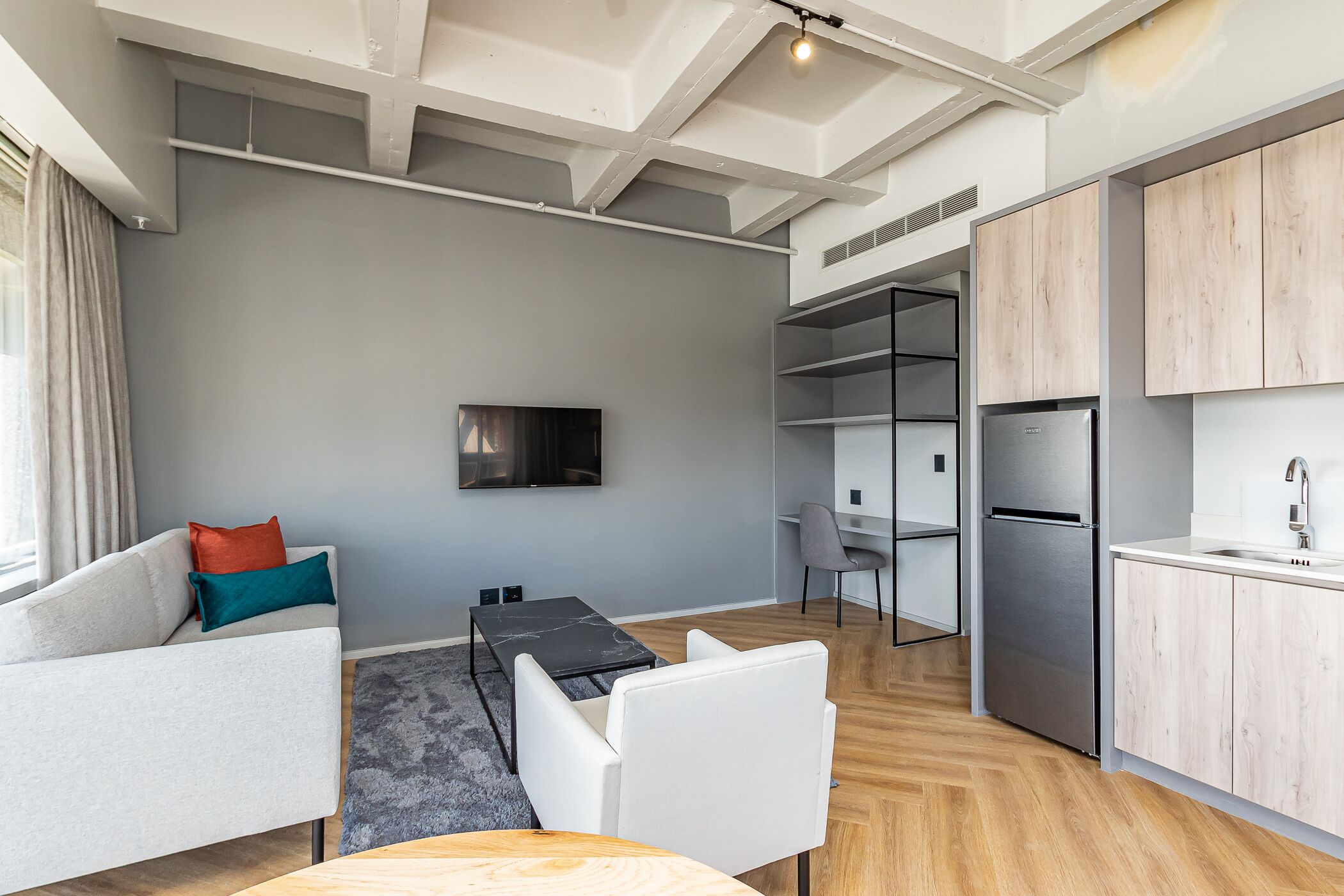 A modern living room with a sofa, a coffee table, and a TV. There is a kitchen area with a refrigerator and a sink.