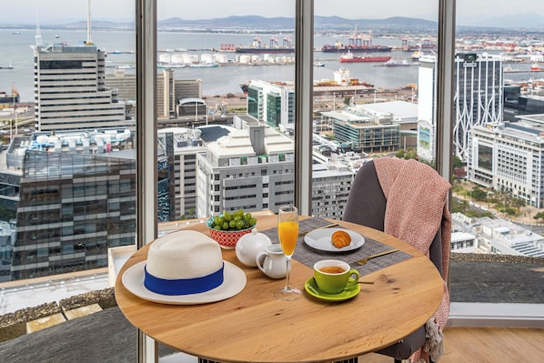 Enjoy a panoramic view of the bustling port city from the comfort of this high-rise room, featuring a spacious seating area and a round table set for breakfast.