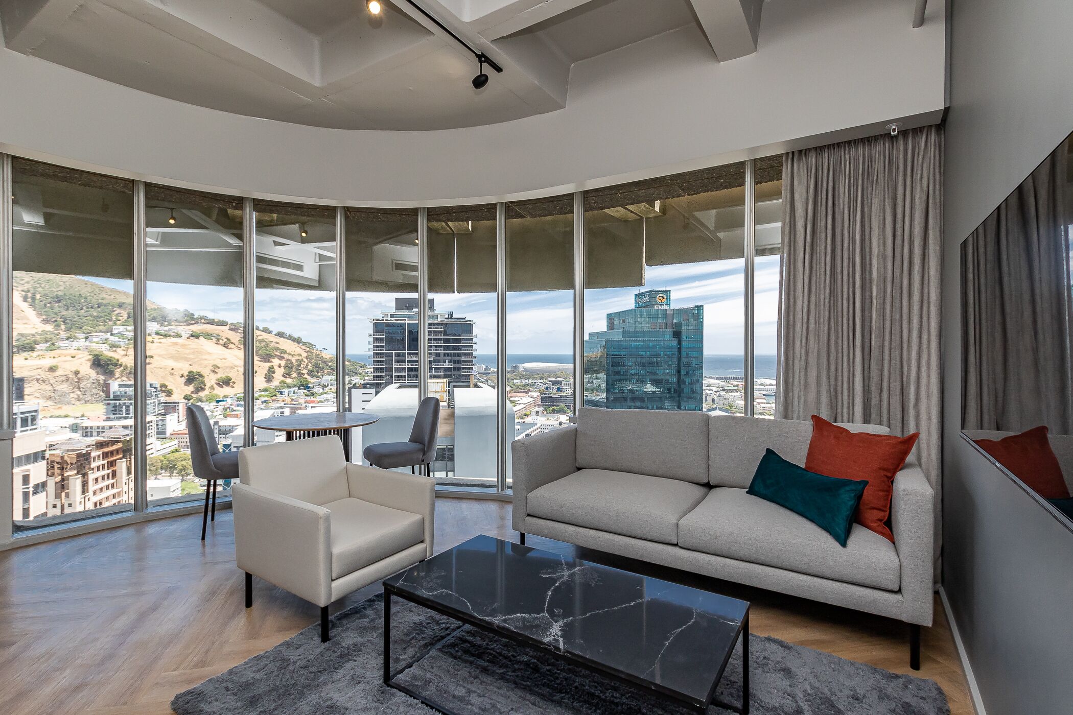 A modern living room with a sofa, armchair, and coffee table, large floor-to-ceiling windows with a city view, and a high ceiling with recessed lighting.