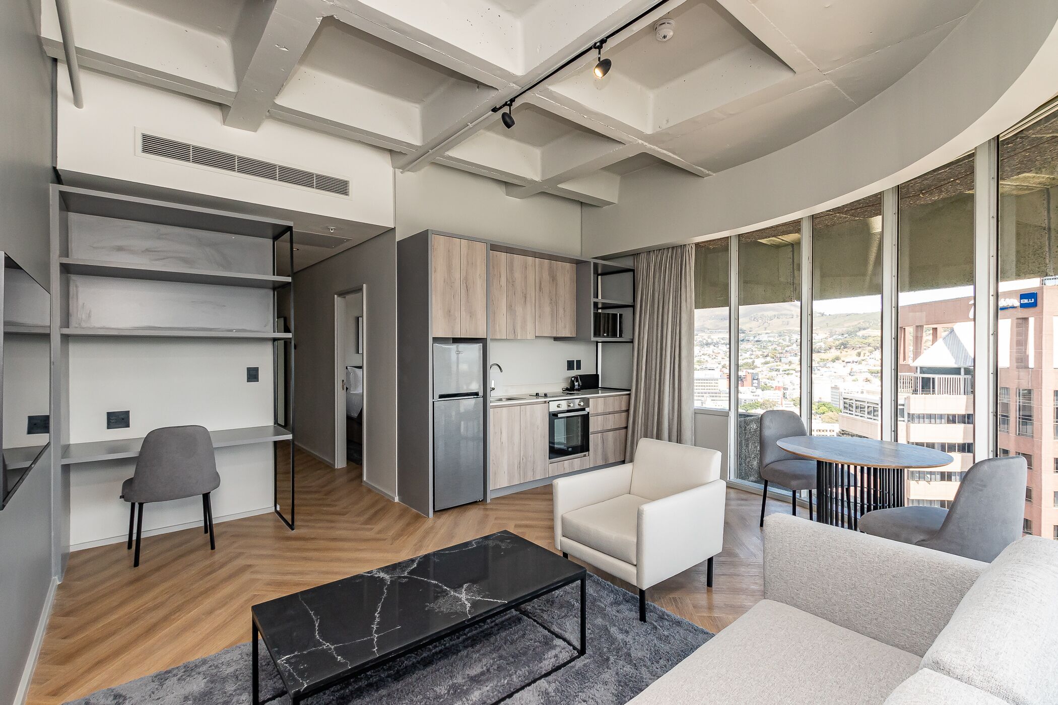 Ein modernes Wohnzimmer mit einer Couch, einem Couchtisch und einem Essbereich mit Tisch und Stühlen. Es gibt eine Küche mit Kühlschrank, Backofen und Mikrowelle. Der Raum verfügt über große Fenster mit Blick auf die Stadt.