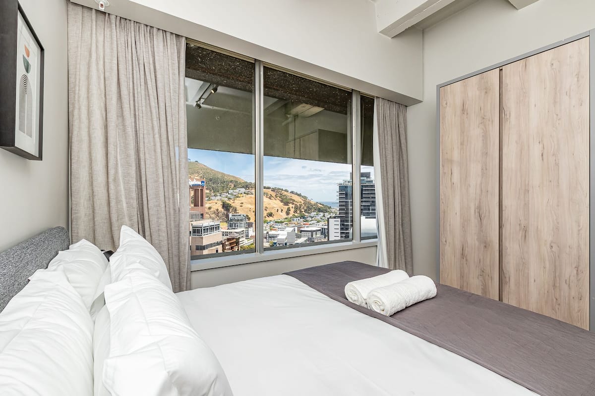 A hotel room with a large window, a bed with white linens, a wooden wardrobe, and a view of a cityscape.