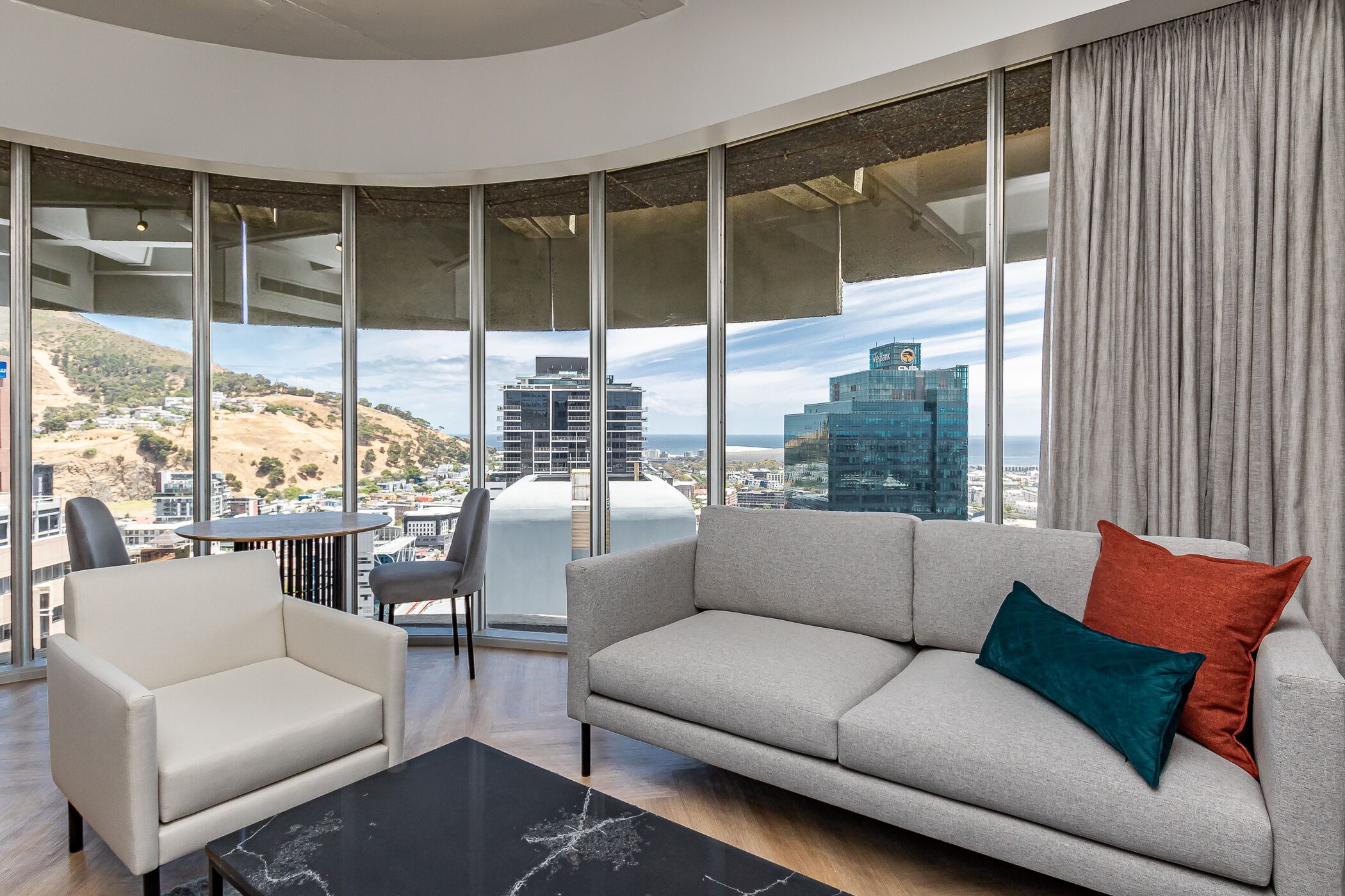 A modern living room with a sofa, armchair, and a dining table. Large windows offer a city view.
