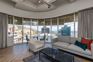 Una sala de estar moderna con sofá, sillón y mesa de centro, ventanas amplias con vista a la ciudad y techo curvo.